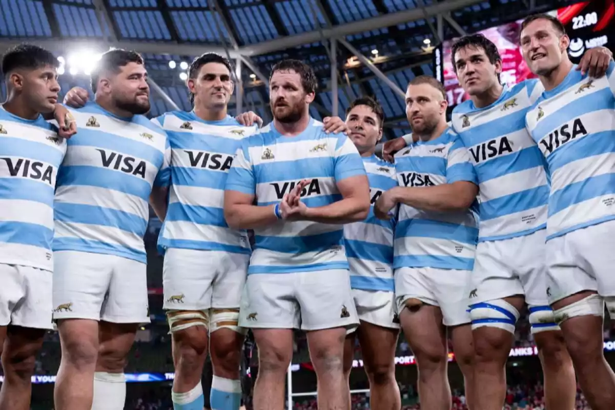 Rugby players in light blue and white uniforms pose together on the field inside a covered stadium.