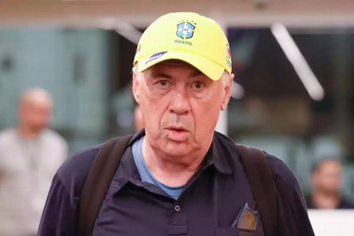Hombre de cabello canoso con gorra amarilla de Brasil y mochila negra mirando al frente en un entorno interior desenfocado Hombre de cabello canoso con gorra amarilla de Brasil y mochila negra mirando al frente en un entorno interior desenfocado
