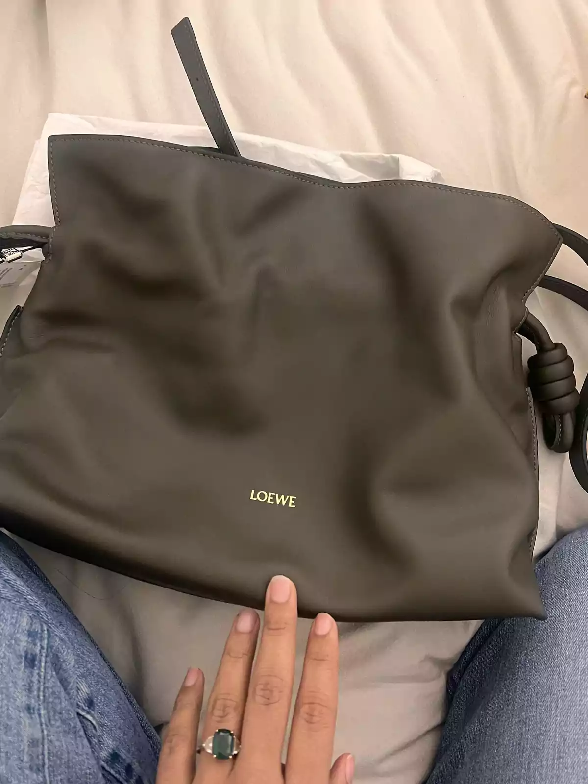 Bolso de cuero color marrón oscuro de la marca Loewe sobre una superficie clara con una mano apoyada en la parte inferior y una persona usando jeans Bolso de cuero color marrón oscuro de la marca Loewe sobre una superficie clara con una mano apoyada en la parte inferior y una persona usando jeans
