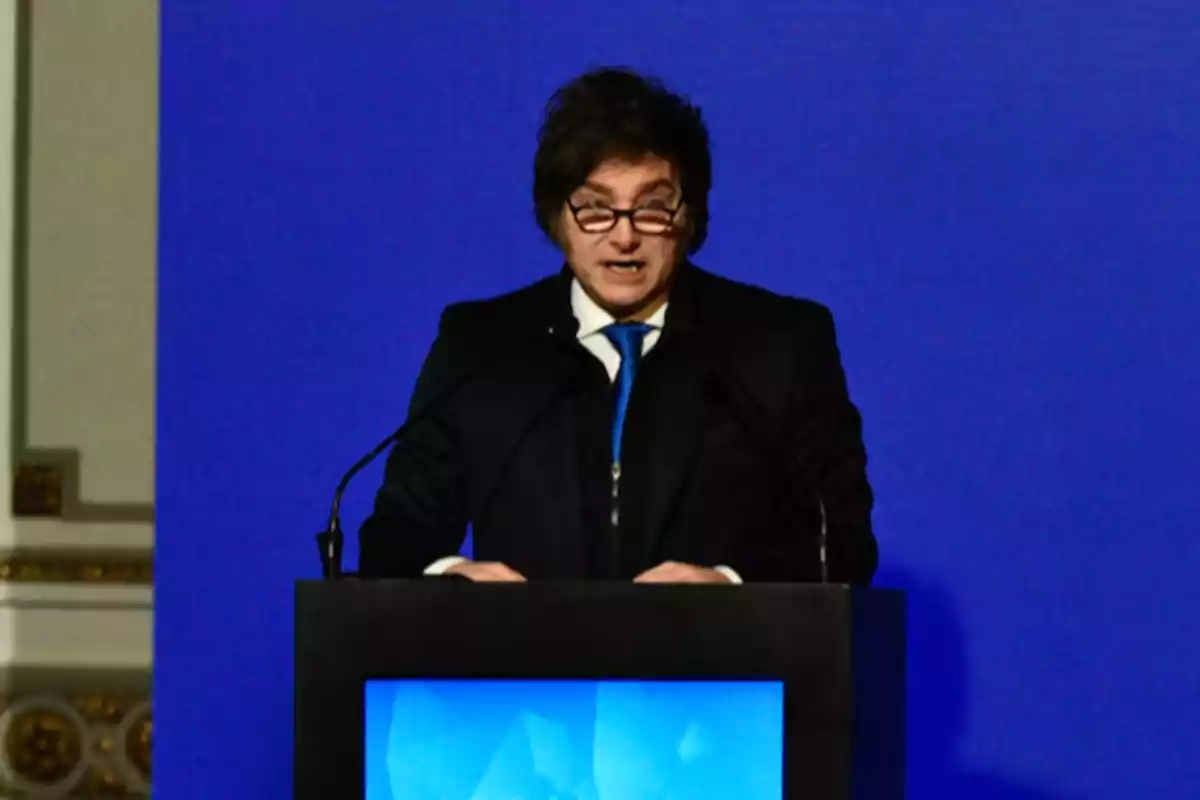 Dark-haired man with glasses speaking at a podium in front of a blue screen Dark-haired man with glasses speaking at a podium in front of a blue screen