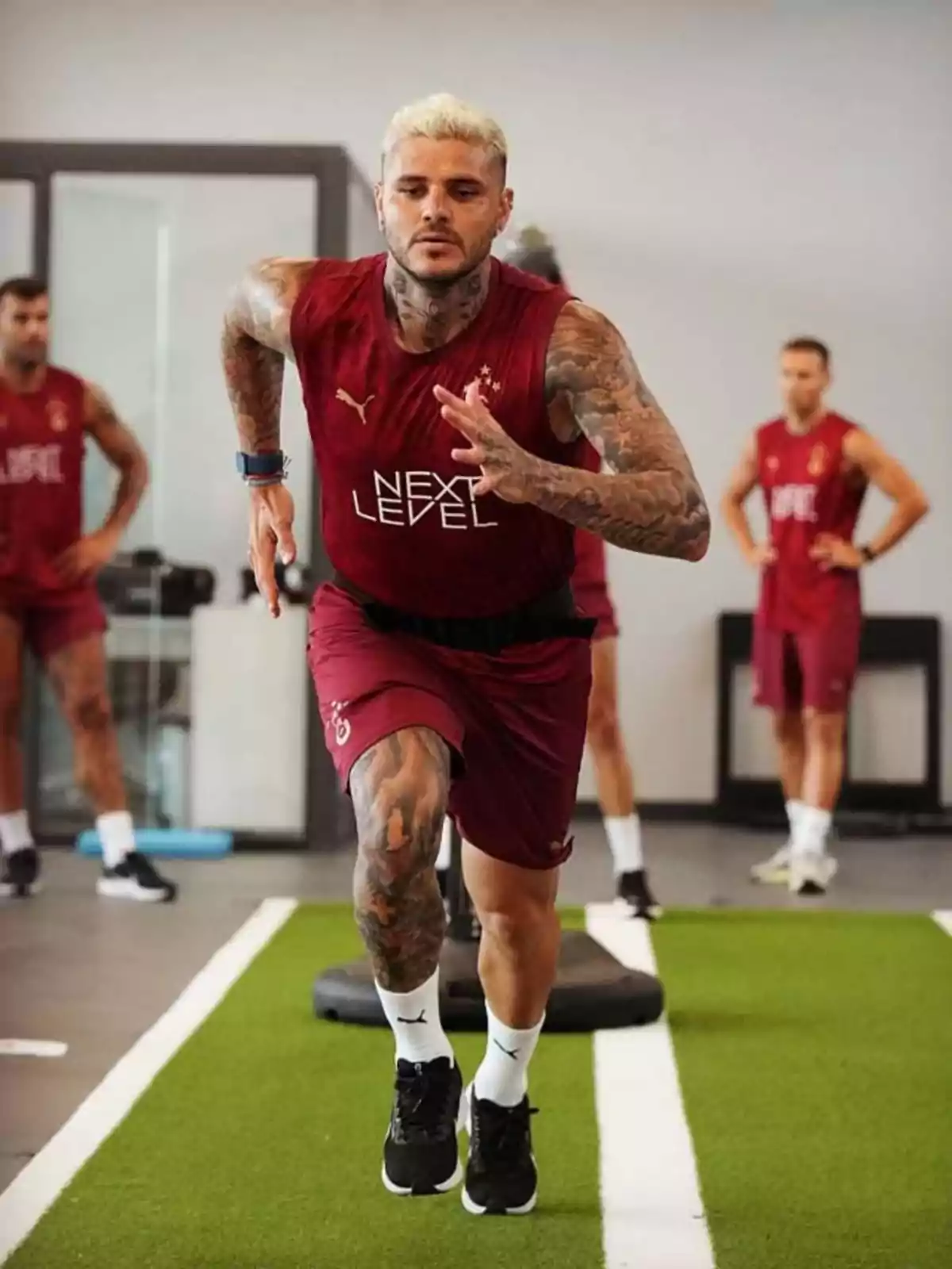 Hombre tatuado con uniforme deportivo rojo realizando un ejercicio de velocidad en un gimnasio sobre una pista de césped artificial mientras otras personas observan al fondo