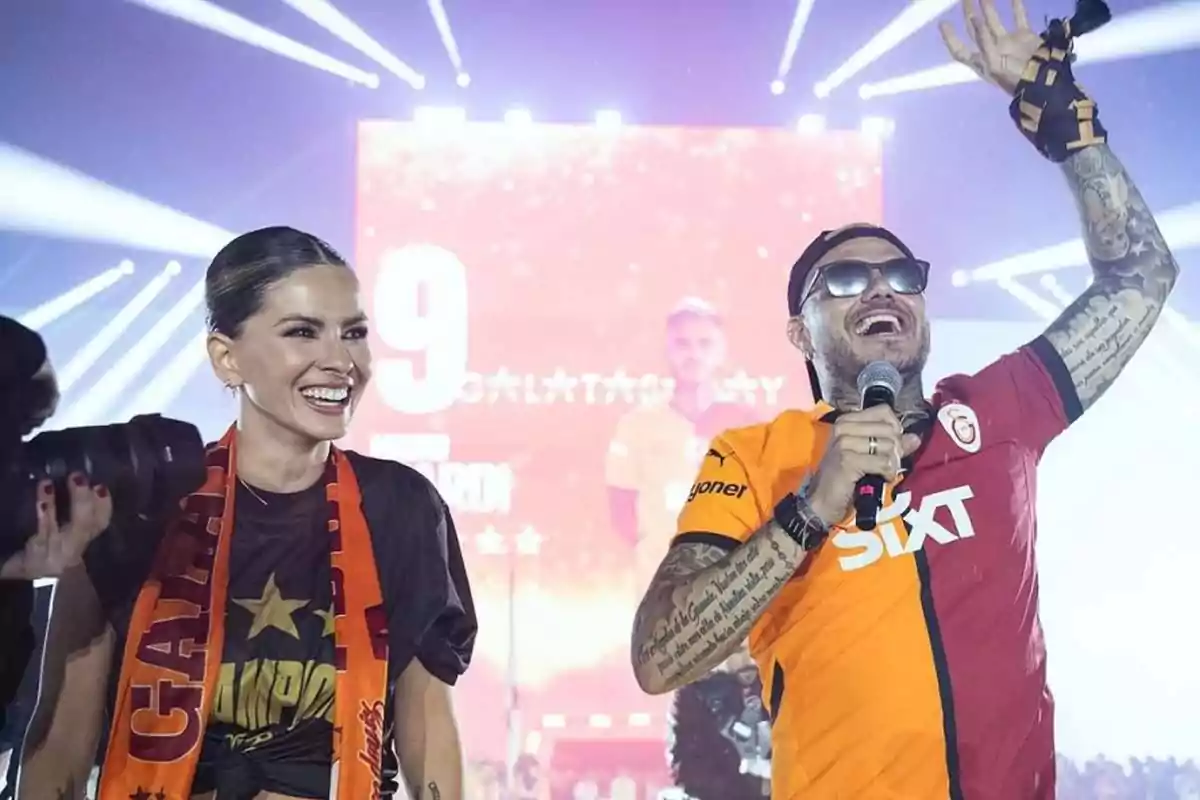 Dos personas celebran en un escenario con luces y una pantalla gigante al fondo, ambos sonríen y llevan camisetas y bufandas del equipo Galatasaray