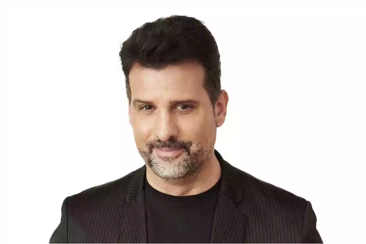 Hombre de cabello oscuro y barba corta con saco negro y camiseta negra sobre fondo blanco