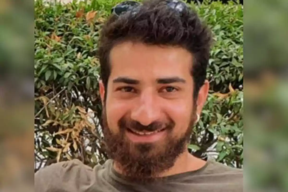Young man with a beard and dark hair smiles in front of a green bush