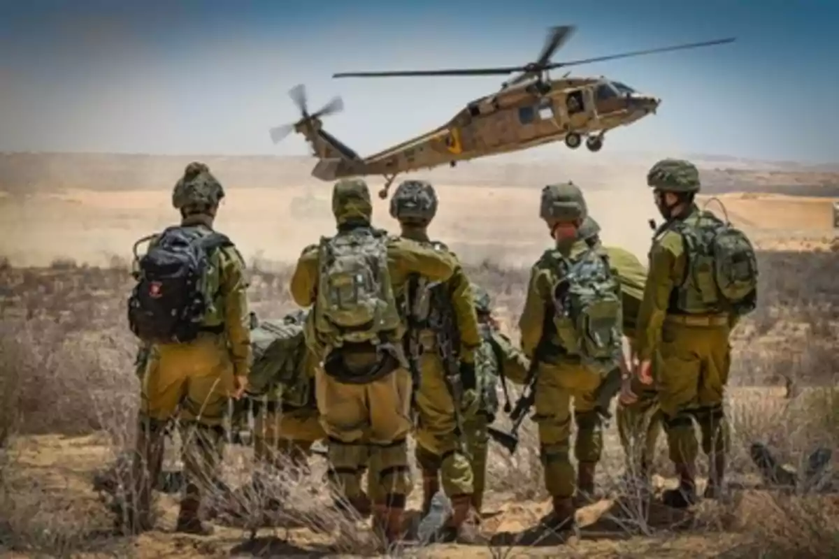 Soldiers with military equipment watch a helicopter landing in a desert area.
