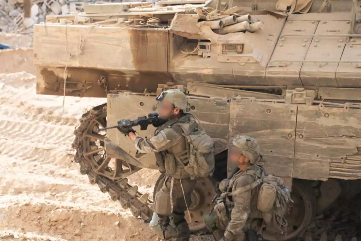 Two soldiers in military uniforms and tactical gear take cover next to a dust-covered armored vehicle on sandy terrain.