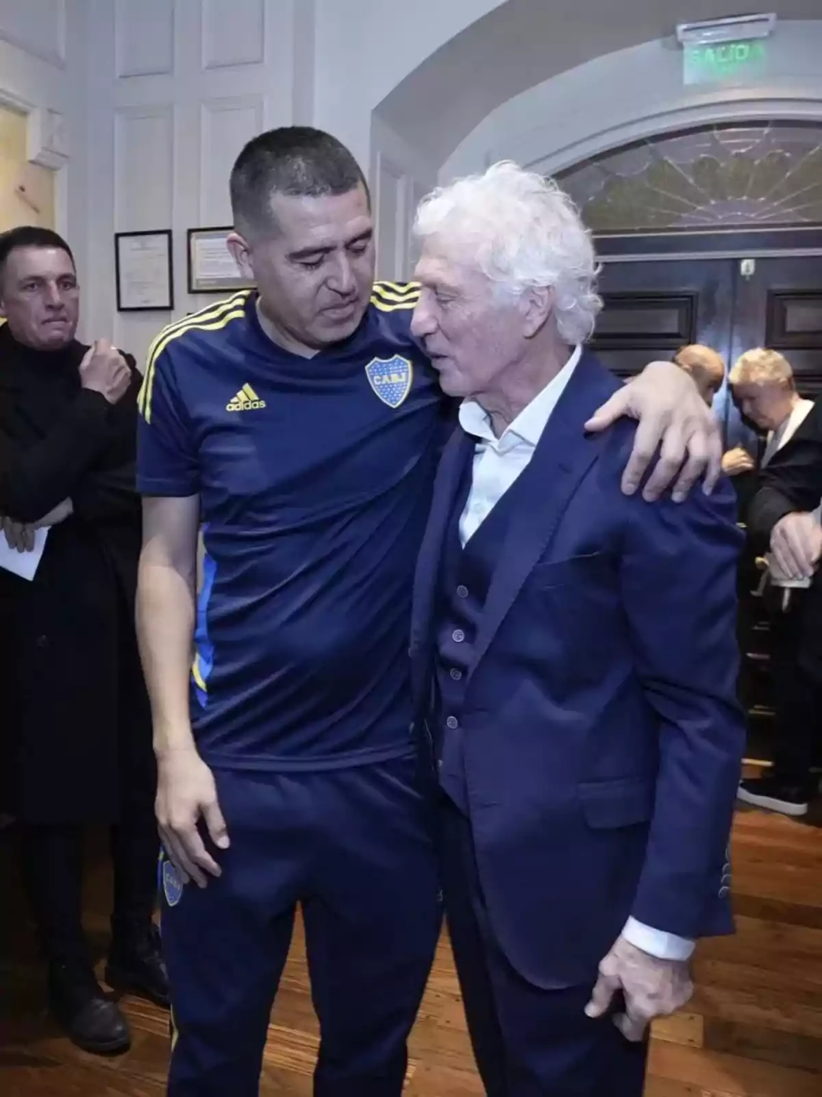 Dos hombres se abrazan y conversan en un ambiente interior elegante, uno lleva ropa deportiva azul con detalles amarillos y el otro viste un traje azul, mientras otras personas observan al fondo.