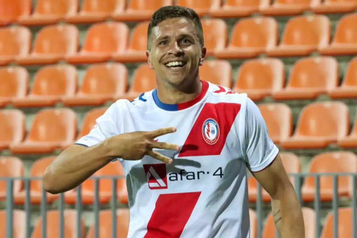 Smiling player celebrating with his hand while wearing a white and red uniform in a stadium with orange stands
