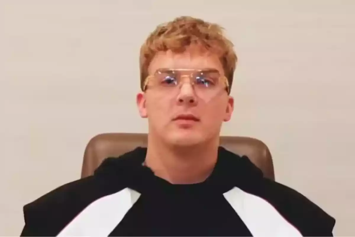 Young person with short blond hair, transparent glasses, and a black sweatjersey sitting in front of a light-colored wall