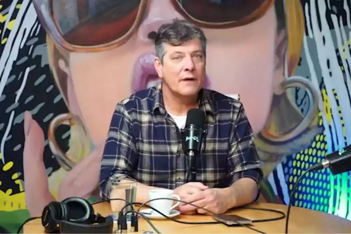 Gray-haired man in a plaid jersey speaking into a microphone in a studio with a colorful mural in the background and recording equipment on the table