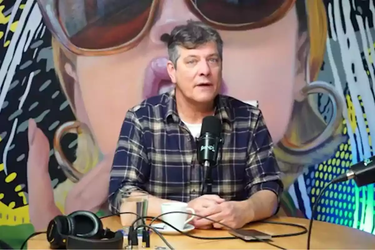 Hombre de cabello canoso y camisa de cuadros hablando frente a un micrófono en un estudio con mural colorido de fondo y audífonos sobre la mesa Hombre de cabello canoso y camisa de cuadros hablando frente a un micrófono en un estudio con mural colorido de fondo y audífonos sobre la mesa