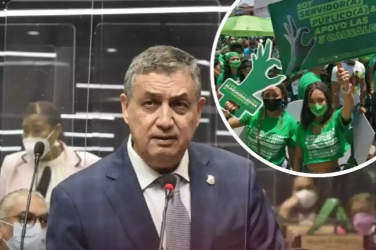 Un hombre de traje habla frente a un micrófono en un recinto oficial mientras en una burbuja al fondo se muestra una manifestación de personas vestidas de verde con pancartas y mascarillas Un hombre de traje habla frente a un micrófono en un recinto oficial mientras en una burbuja al fondo se muestra una manifestación de personas vestidas de verde con pancartas y mascarillas