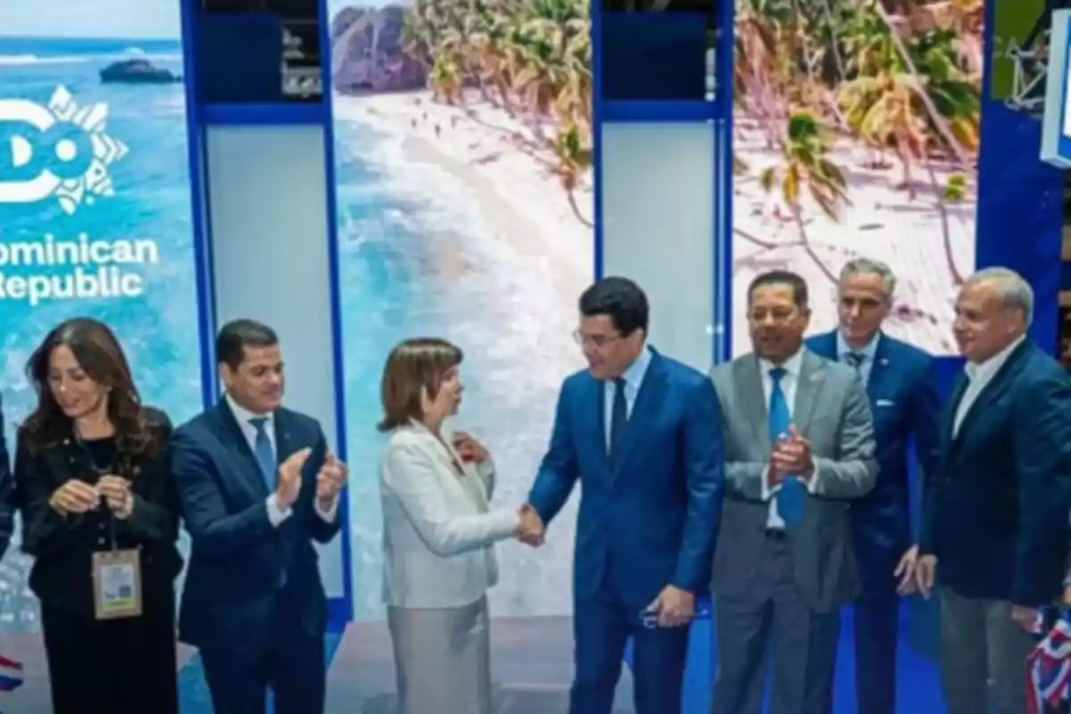Varias personas vestidas de manera formal se encuentran en un evento frente a un fondo que muestra una playa tropical y el texto Dominican Republic, dos de ellas se dan la mano mientras los demás observan y aplauden.