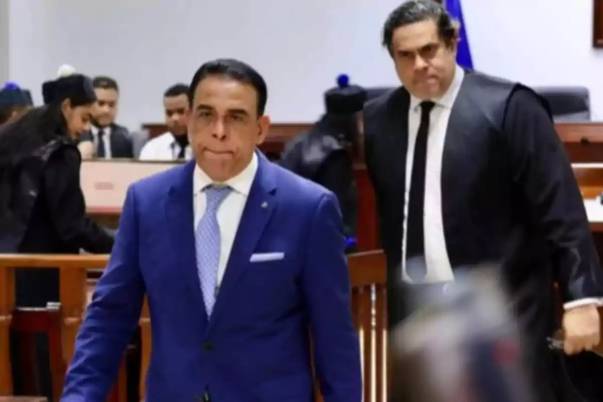 Dos hombres con expresión seria en una sala de tribunal, uno de ellos lleva traje azul y corbata clara, el otro viste toga negra y corbata, al fondo se observan más personas en ambiente judicial. Dos hombres con expresión seria en una sala de tribunal, uno de ellos lleva traje azul y corbata clara, el otro viste toga negra y corbata, al fondo se observan más personas en ambiente judicial.