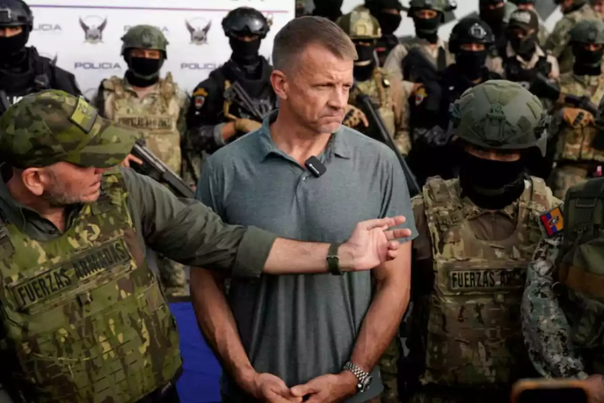 Un hombre de camiseta gris es escoltado por militares con uniforme de camuflaje y chalecos antibalas mientras otros soldados armados observan al fondo Un hombre de camiseta gris es escoltado por militares con uniforme de camuflaje y chalecos antibalas mientras otros soldados armados observan al fondo