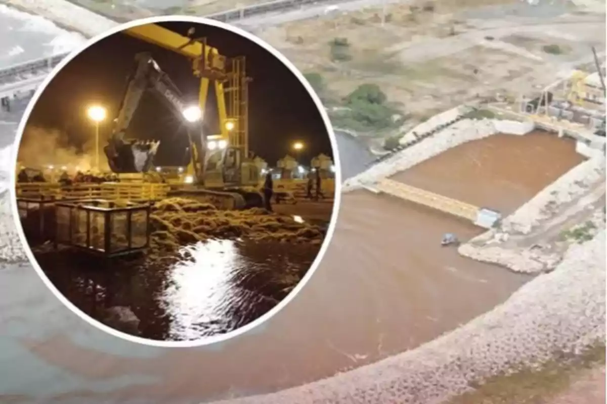 Excavadora trabajando de noche junto a un río con agua marrón y una estructura de contención en una zona de construcción