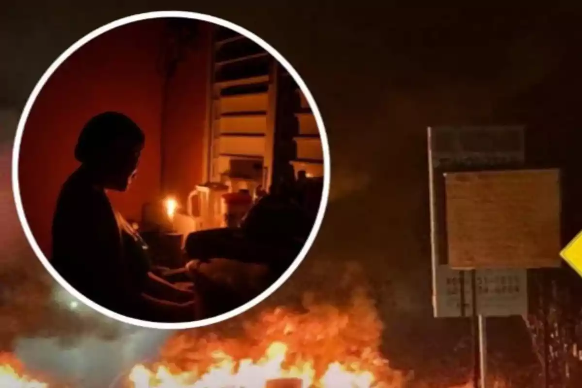Una mujer sentada a oscuras con una vela encendida y al fondo una calle con fuego y humo