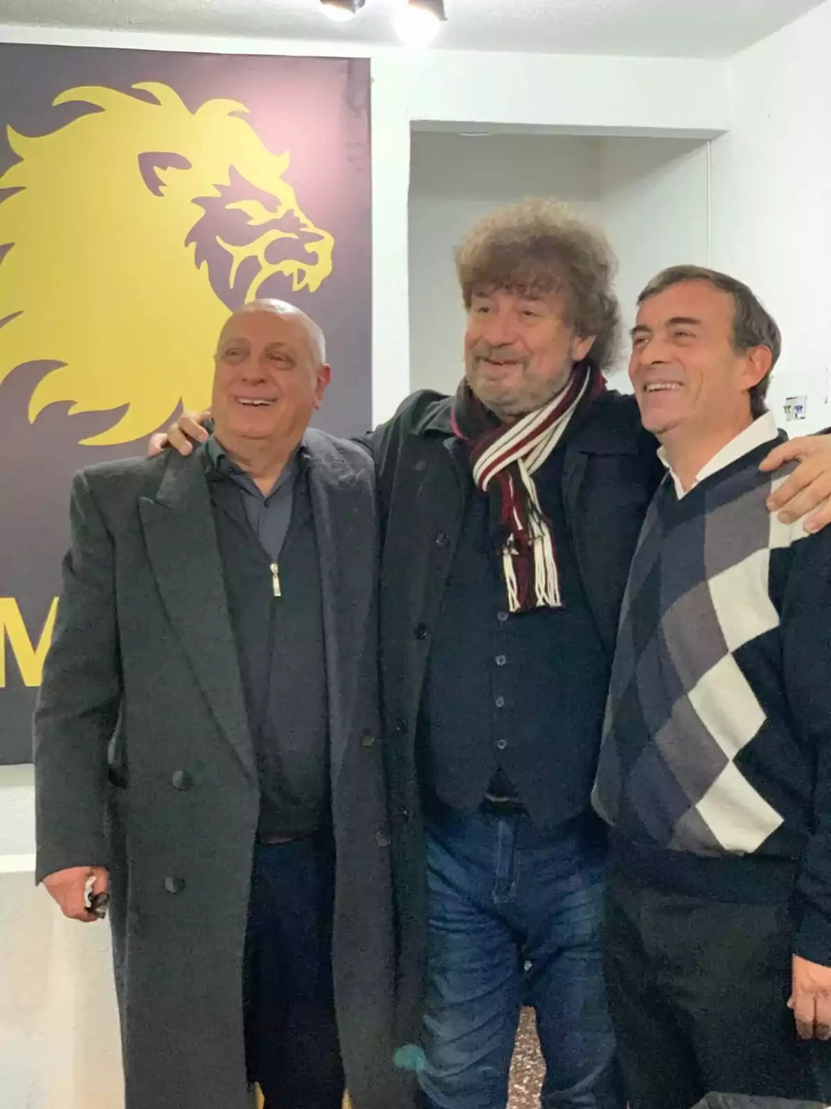 Tres hombres sonríen y posan juntos frente a una pared blanca con un cartel de un león amarillo Tres hombres sonríen y posan juntos frente a una pared blanca con un cartel de un león amarillo