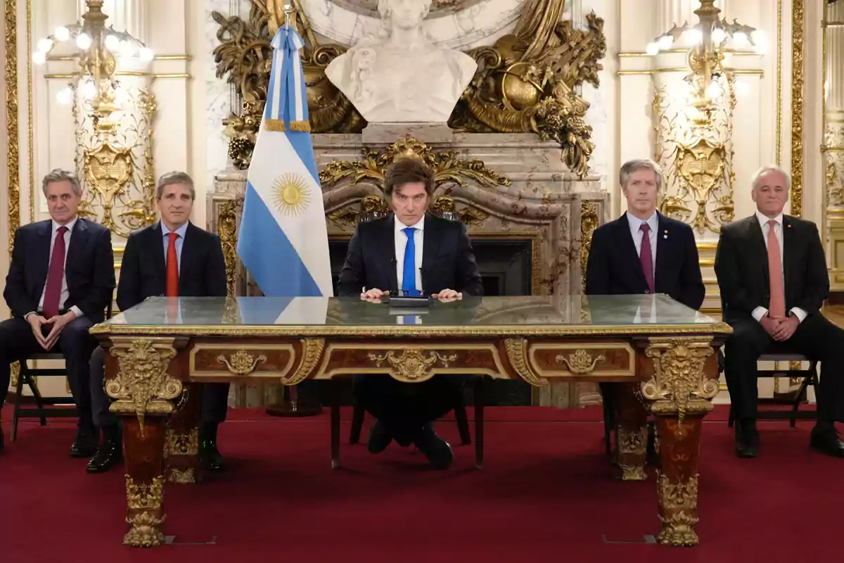 Cinco hombres con traje sentados en una sala elegante con decoración dorada y una bandera de Argentina detrás de una mesa de madera tallada