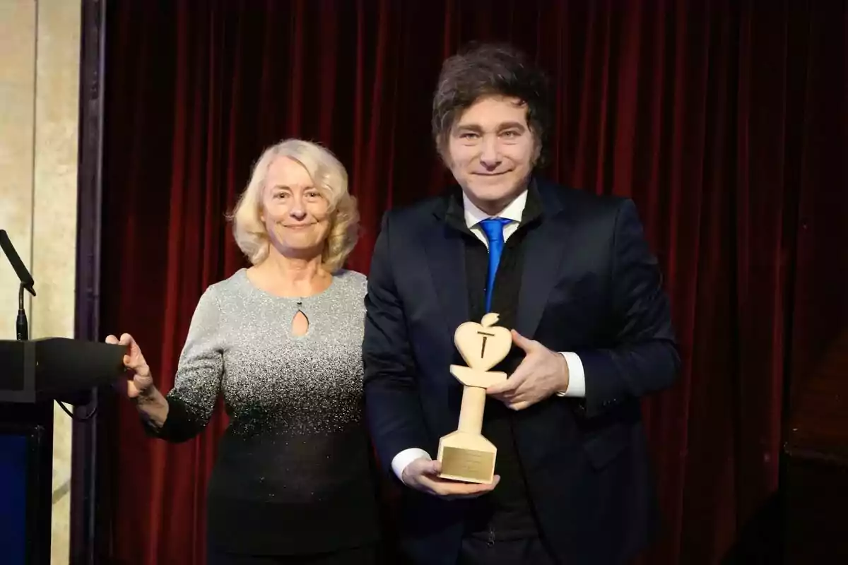 Two people pose in front of a red curtain, one of them holding a wooden apple-shaped trophy.
