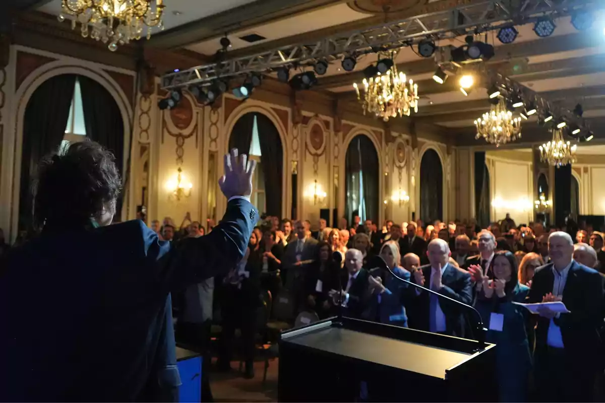 Person seen from behind waving to an audience applauding in an elegant event hall with chandeliers and classic decor Person seen from behind waving to an audience applauding in an elegant event hall with chandeliers and classic decor