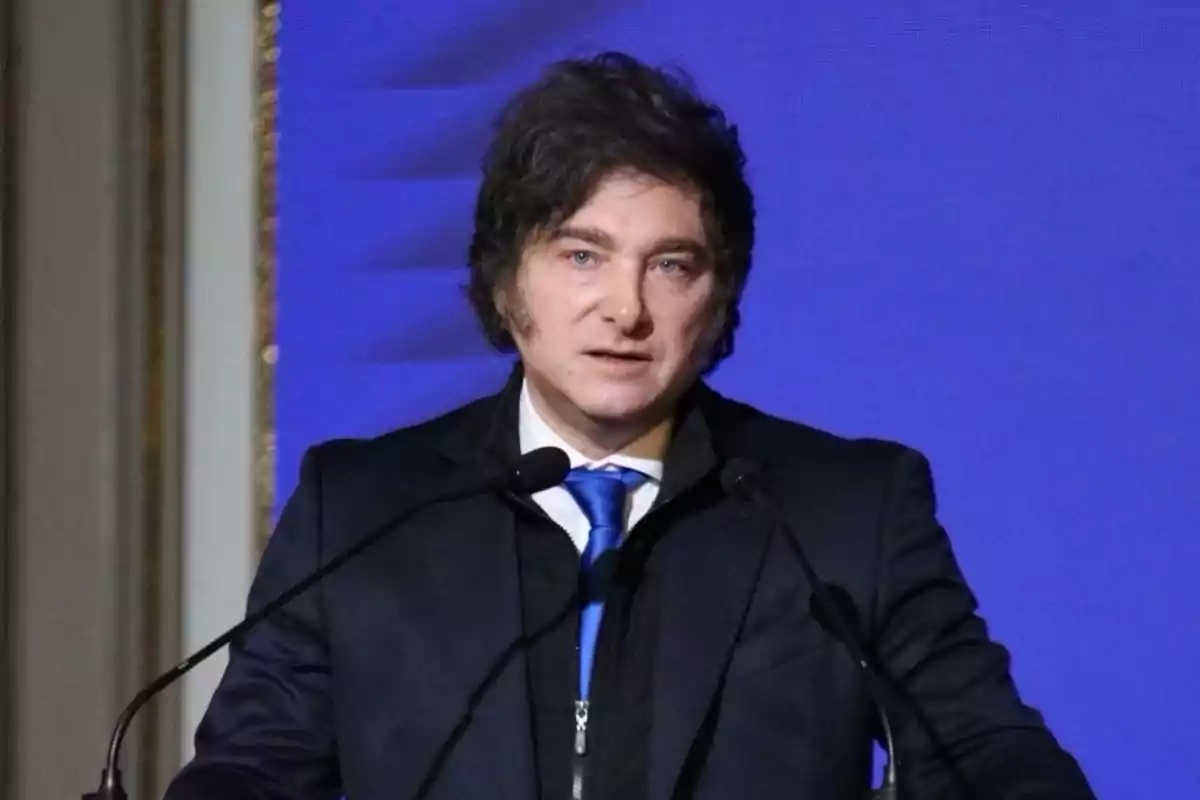 Dark-haired man in a blue suit speaking in front of two microphones with a blue background