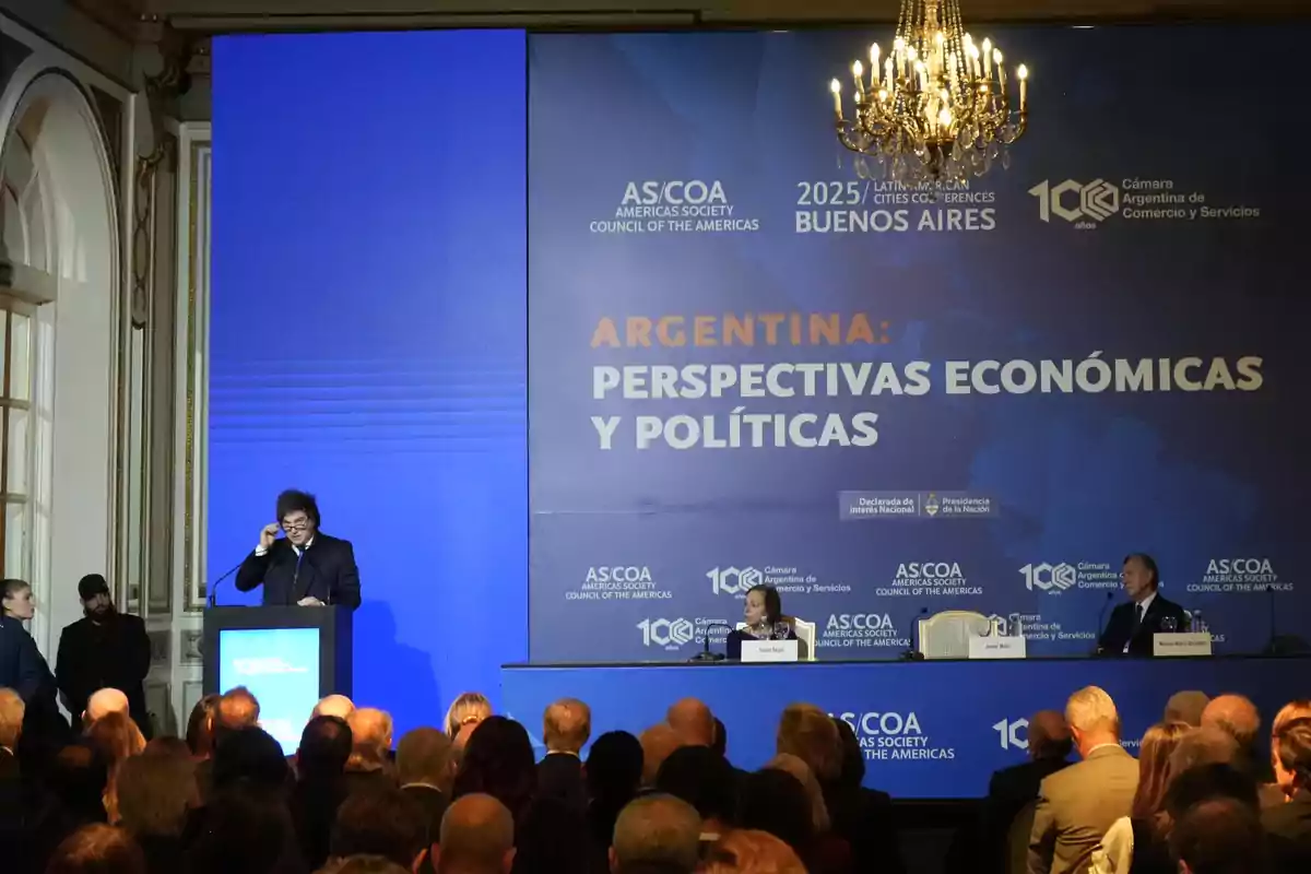 A man in a suit speaks at a podium in front of an audience at an event titled Argentina Economic and Political Perspectives, with a large blue banner in the background and a chandelier hanging from the ceiling. A man in a suit speaks at a podium in front of an audience at an event titled Argentina Economic and Political Perspectives, with a large blue banner in the background and a chandelier hanging from the ceiling.