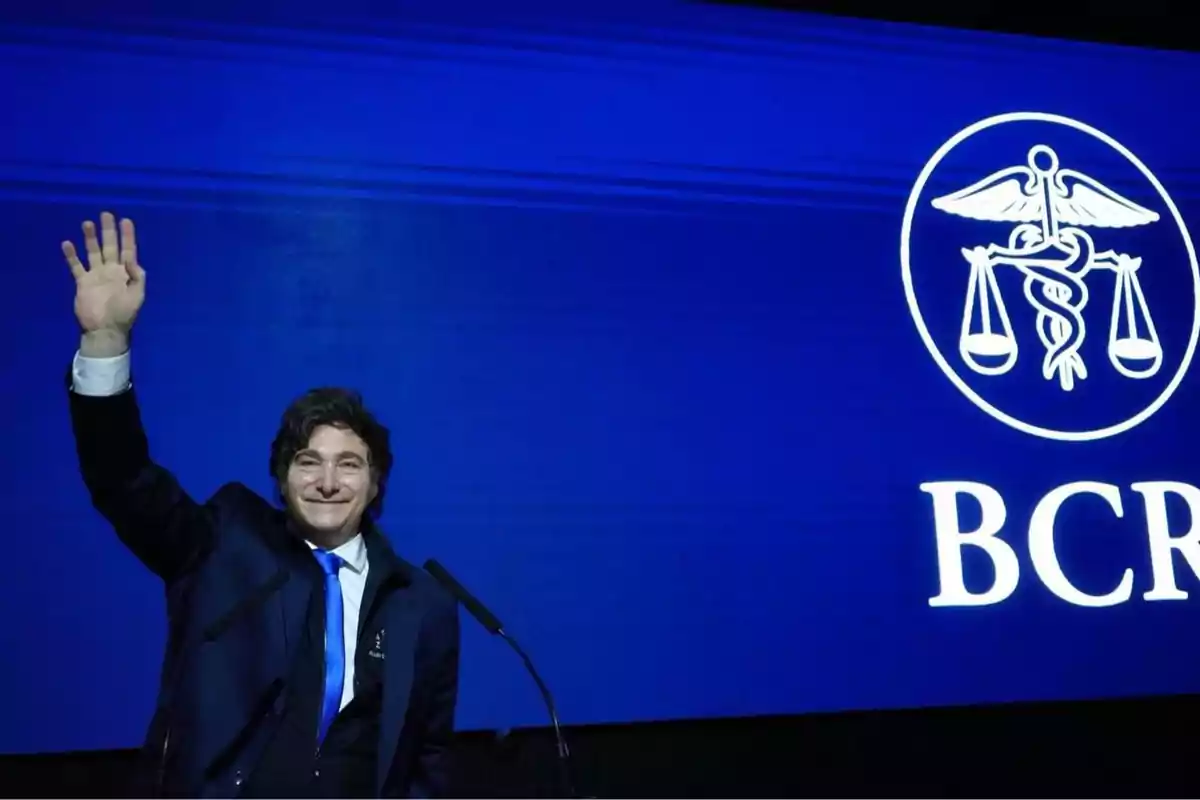 Hombre de traje oscuro y corbata azul saludando con la mano levantada frente a una pantalla azul con el logo y las letras BCRA