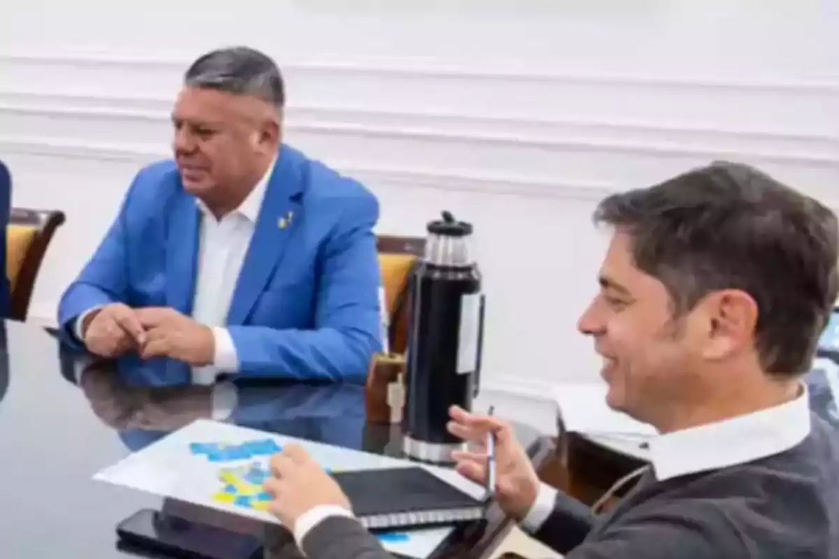Dos hombres sentados en una mesa de reuniones conversan mientras uno de ellos sostiene un bolígrafo y una libreta, con un termo y un mate sobre la mesa Dos hombres sentados en una mesa de reuniones conversan mientras uno de ellos sostiene un bolígrafo y una libreta, con un termo y un mate sobre la mesa