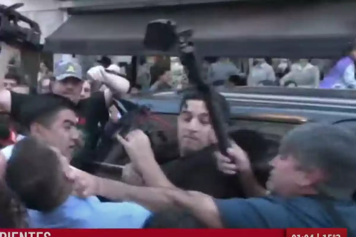 Several people struggle and push each other in the street while some hold cameras and microphones amid a chaotic situation in front of a vehicle. Several people struggle and push each other in the street while some hold cameras and microphones amid a chaotic situation in front of a vehicle.