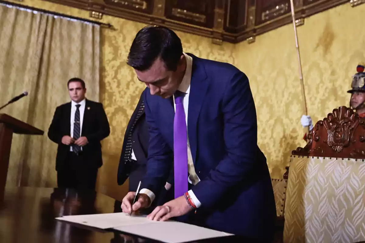 Man in a blue suit and purple tie signing a document in an elegant room with golden walls, while a guard and another person watch in the background.