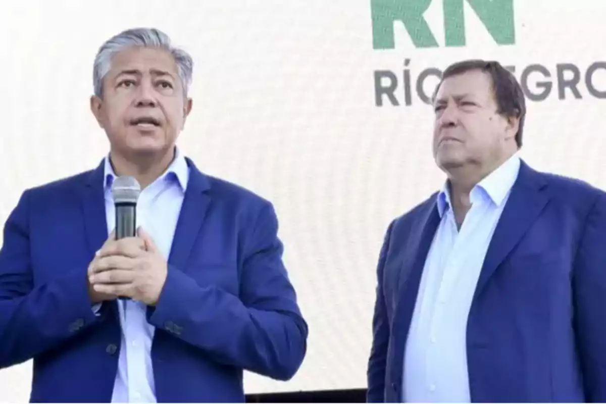 Two men in blue suits are standing side by side, one of them is holding a microphone, and in the background there is a sign with the word Río Negro. Two men in blue suits are standing side by side, one of them is holding a microphone, and in the background there is a sign with the word Río Negro.