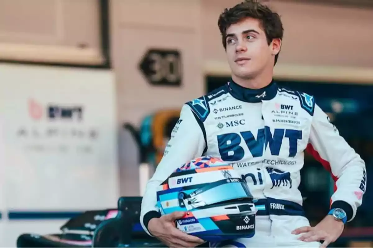 Motorsport driver in a white and blue racing suit holding a helmet while sitting in a Formula 1 team paddock