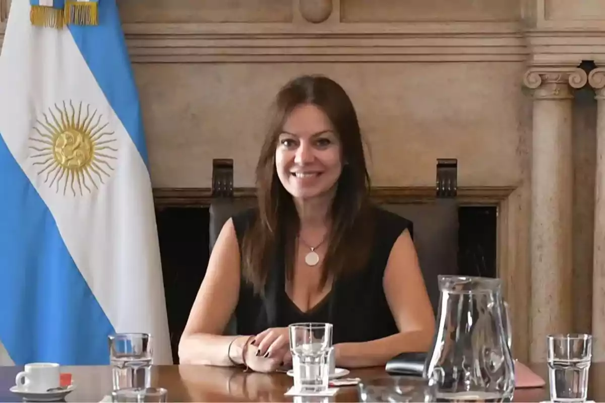 Mujer sentada en una mesa de reuniones con una bandera de Argentina al fondo y varios vasos de agua sobre la mesa
