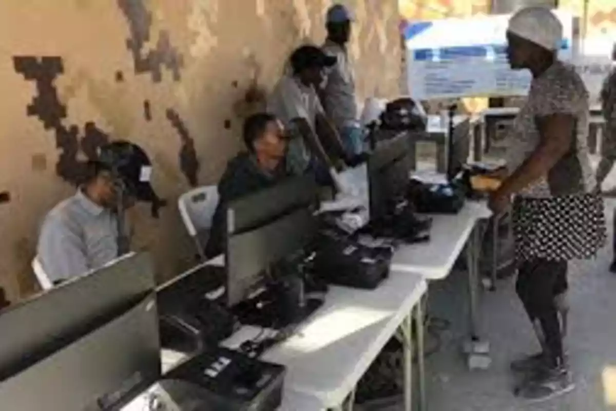 Varias personas trabajan en una mesa con computadoras y equipos electrónicos mientras una mujer se acerca para ser atendida en un espacio al aire libre Varias personas trabajan en una mesa con computadoras y equipos electrónicos mientras una mujer se acerca para ser atendida en un espacio al aire libre