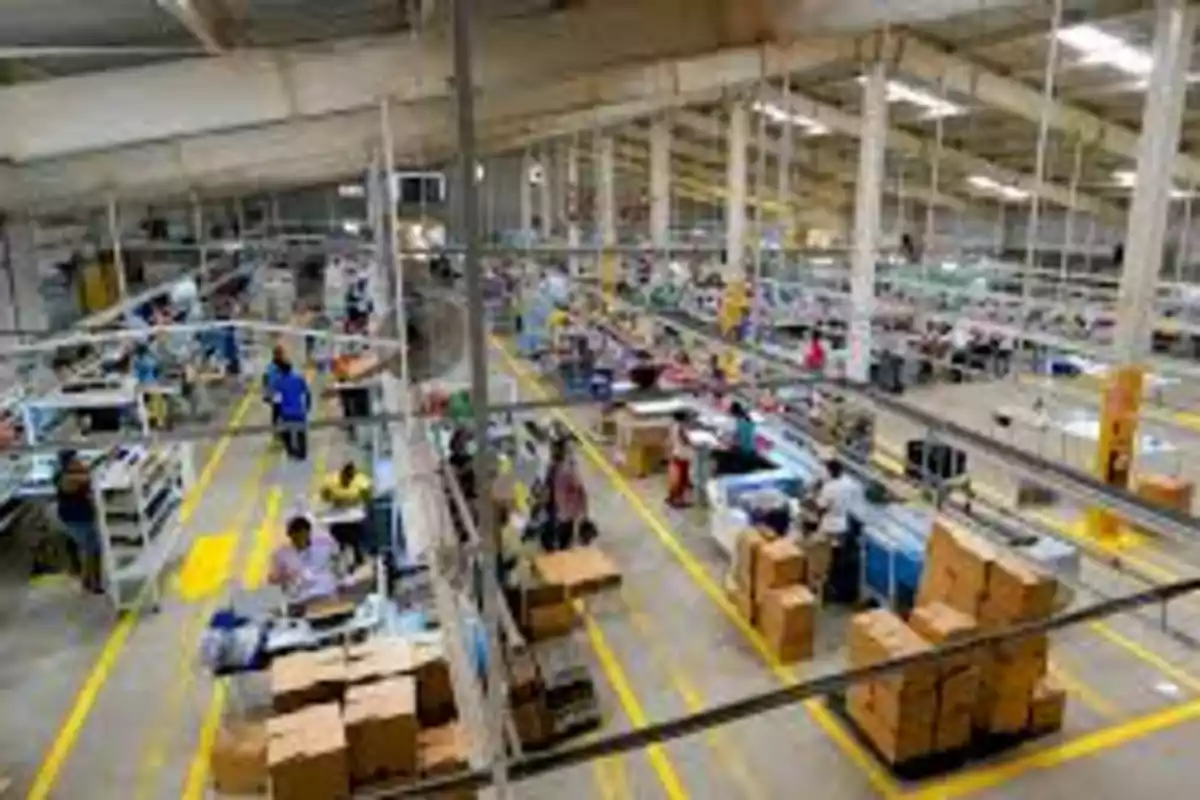 Vista general de una fábrica con varias personas trabajando en diferentes estaciones y cajas apiladas en el suelo Vista general de una fábrica con varias personas trabajando en diferentes estaciones y cajas apiladas en el suelo