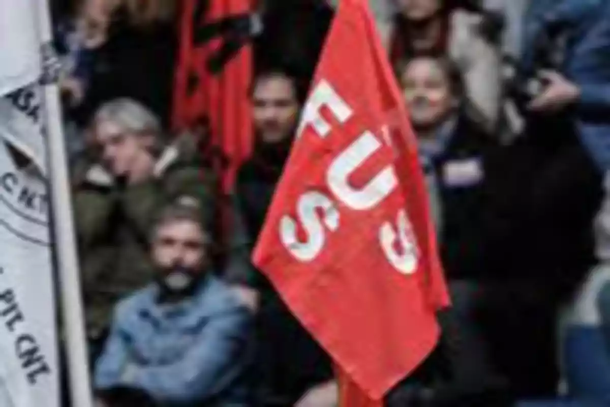 Bandera roja con letras blancas sostenida frente a un grupo de personas desenfocadas en lo que parece ser una manifestación o reunión. Bandera roja con letras blancas sostenida frente a un grupo de personas desenfocadas en lo que parece ser una manifestación o reunión.