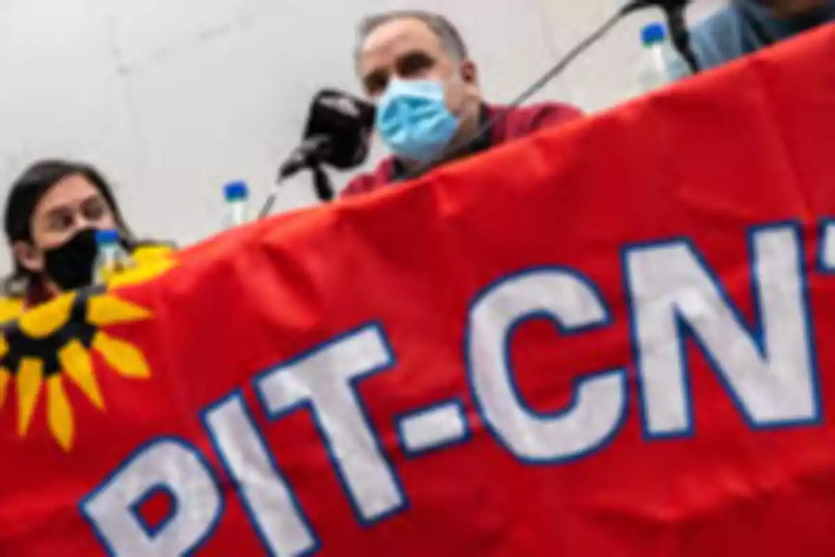 Personas con tapabocas sentadas detrás de una mesa cubierta con una bandera roja de PIT-CNT Personas con tapabocas sentadas detrás de una mesa cubierta con una bandera roja de PIT-CNT