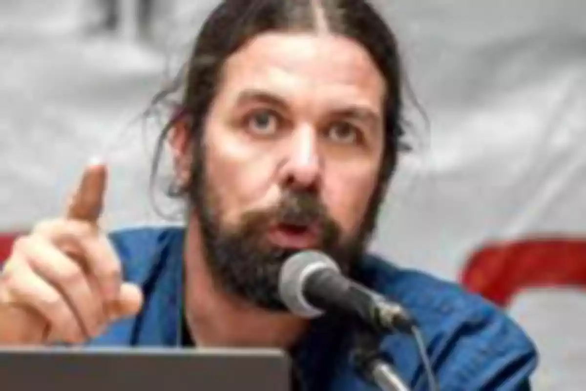 Hombre de cabello largo y barba hablando frente a un micrófono mientras levanta un dedo Hombre de cabello largo y barba hablando frente a un micrófono mientras levanta un dedo