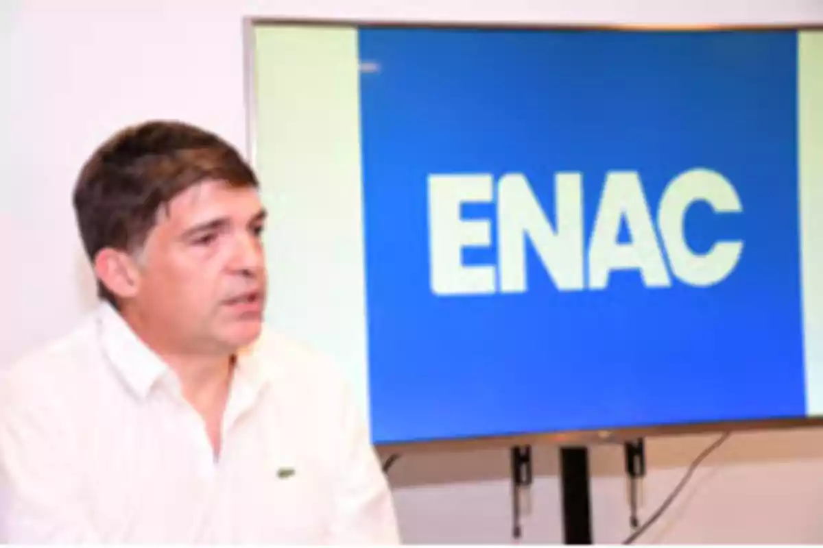 Short-haired man in a white jersey sitting in front of a blue screen with the word ENAC in large letters. Short-haired man in a white jersey sitting in front of a blue screen with the word ENAC in large letters.