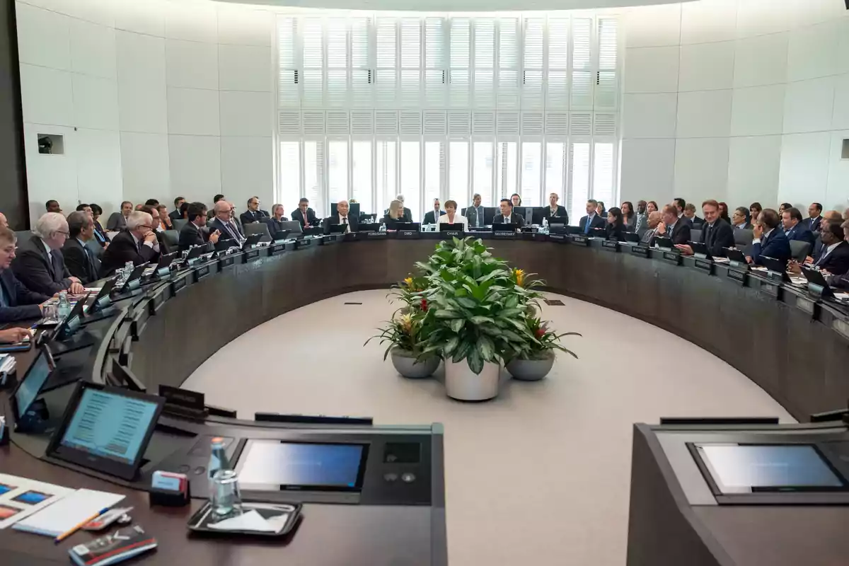 Personas reunidas en una sala de conferencias moderna sentadas alrededor de una mesa circular con plantas en el centro y pantallas frente a cada participante Personas reunidas en una sala de conferencias moderna sentadas alrededor de una mesa circular con plantas en el centro y pantallas frente a cada participante