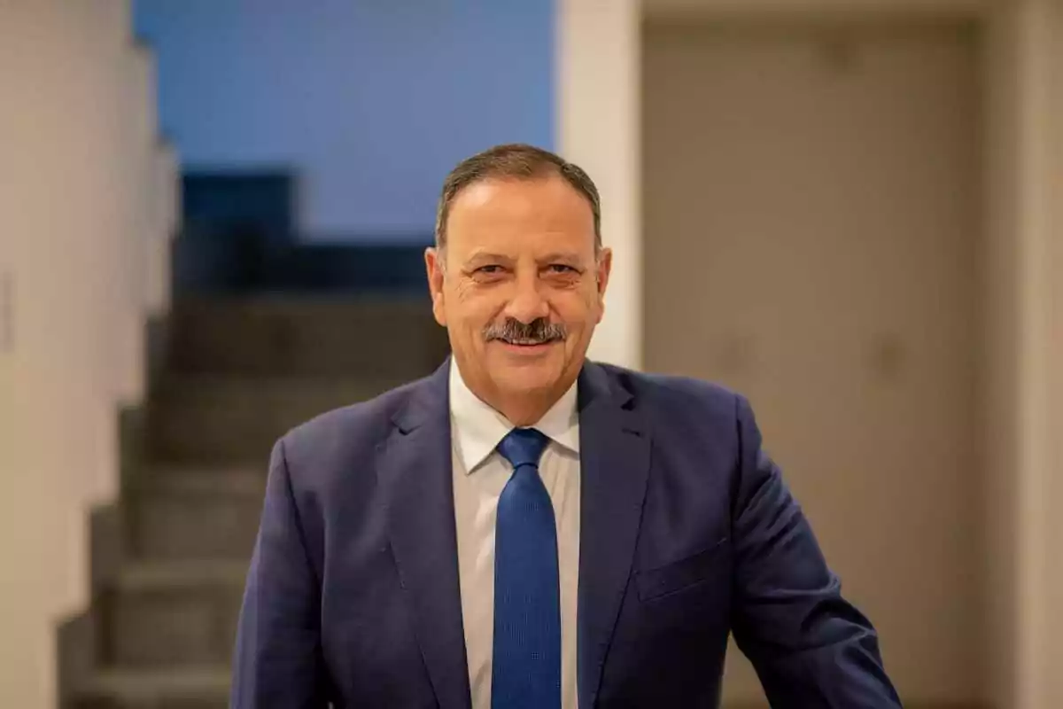 Hombre de mediana edad con bigote y cabello corto, vestido con traje azul y corbata, sonriendo en un entorno interior con escaleras al fondo Hombre de mediana edad con bigote y cabello corto, vestido con traje azul y corbata, sonriendo en un entorno interior con escaleras al fondo