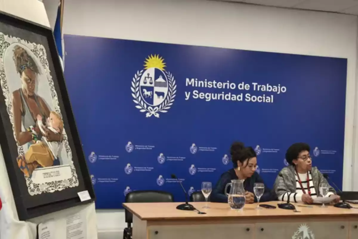 Dos mujeres sentadas en una mesa durante una conferencia de prensa en el Ministerio de Trabajo y Seguridad Social de Uruguay, con un cuadro de una mujer amamantando a un bebé en primer plano. Dos mujeres sentadas en una mesa durante una conferencia de prensa en el Ministerio de Trabajo y Seguridad Social de Uruguay, con un cuadro de una mujer amamantando a un bebé en primer plano.