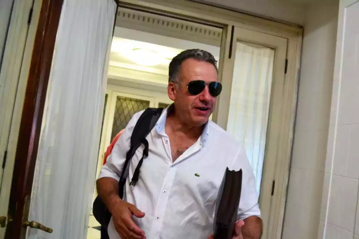 Hombre de mediana edad con camisa blanca y gafas de sol entrando a una habitación mientras lleva una mochila y una carpeta en la mano Hombre de mediana edad con camisa blanca y gafas de sol entrando a una habitación mientras lleva una mochila y una carpeta en la mano