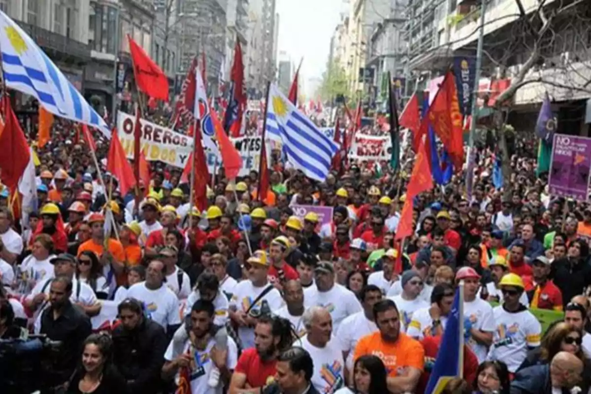 Manifestación multitudinaria en una ciudad con personas portando banderas de Uruguay, pancartas y cascos de seguridad Manifestación multitudinaria en una ciudad con personas portando banderas de Uruguay, pancartas y cascos de seguridad