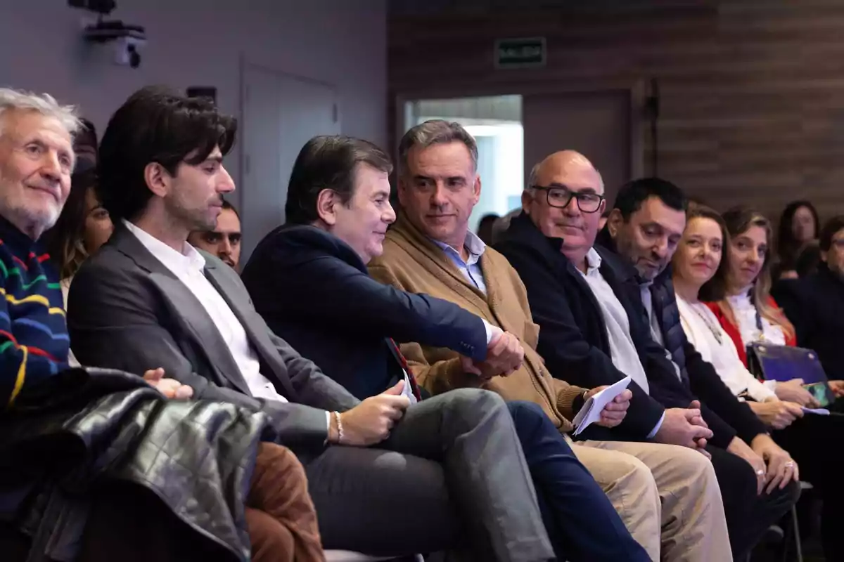 Un grupo de personas sentadas en una fila durante un evento, dos de ellas se dan la mano mientras conversan y el resto observa atentamente Un grupo de personas sentadas en una fila durante un evento, dos de ellas se dan la mano mientras conversan y el resto observa atentamente