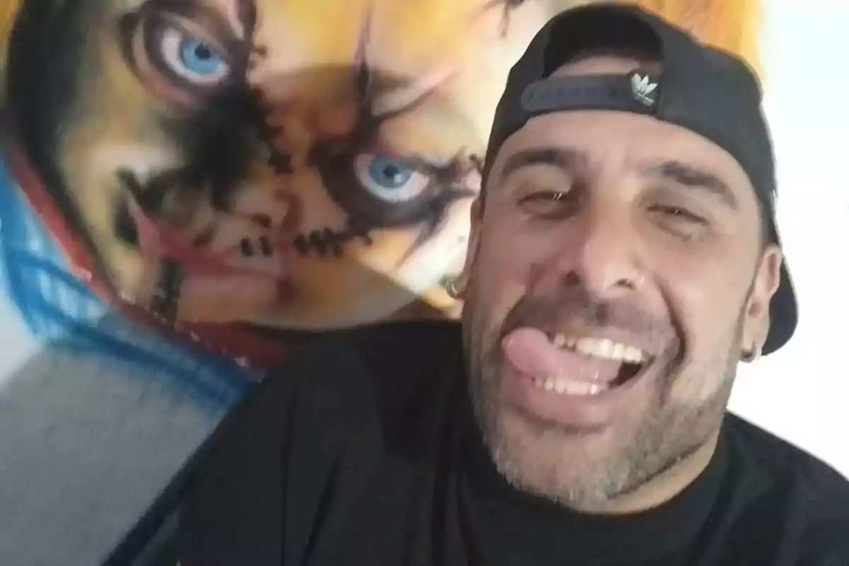 Hombre con gorra negra sacando la lengua mientras se toma una selfie frente a un mural del personaje Chucky
