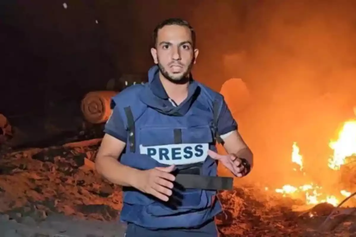 Journalist wearing a bulletproof vest labeled PRESS reporting in front of a fire at night