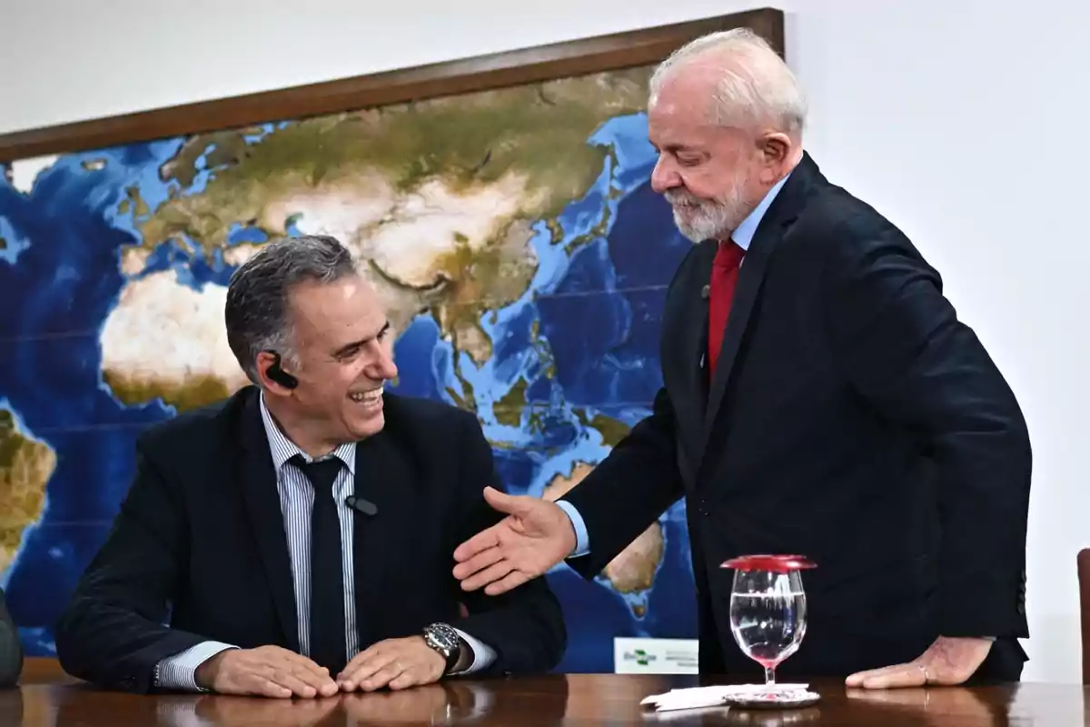 Dos hombres de traje interactúan sonrientes en una sala de reuniones con un mapa del mundo de fondo y una copa de agua sobre la mesa