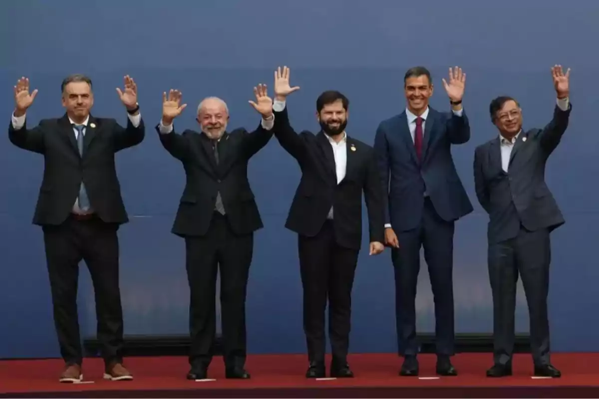 Cinco hombres con traje y corbata posan de pie sobre una alfombra roja frente a un fondo azul, levantando la mano en señal de saludo. Cinco hombres con traje y corbata posan de pie sobre una alfombra roja frente a un fondo azul, levantando la mano en señal de saludo.